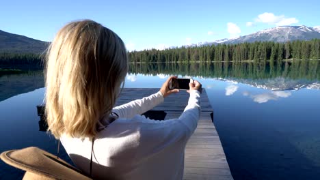 Young-woman-standing-on-wooden-pier-above-lake-takes-a-photo-of-stunning-mountain-lake-scenery-using-her-mobile-phone