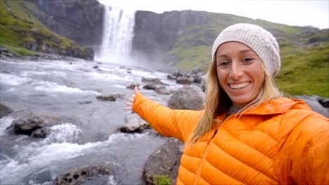 Young-woman-taking-selfie-portrait-with-magnificent-waterfall-in-Iceland,-blowing-a-kiss-to-camera.-People-travel-exploration-concept