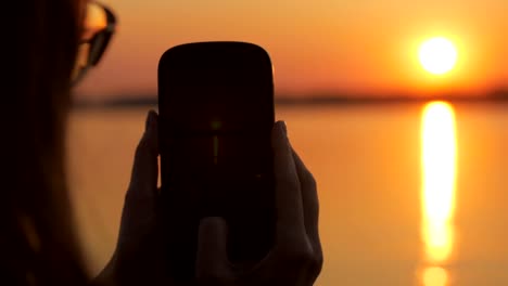 woman-is-taking-photo-of-a-beautiful-vivid-sunset-over-river,-using-her-smartphone,-close-up