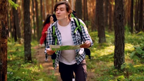 Smiling-young-man-is-looking-at-map-and-searching-for-right-way-in-forest-while-multiethnic-group-of-friends-is-following-him.-Friendship,-nature-and-people-concept.