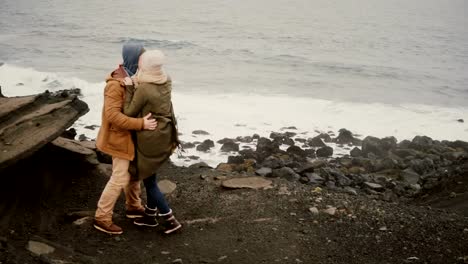 Back-view-of-young-man-and-woman-standing-on-the-shore-of-the-sea-in-cold-day,-holding-hands-and-looking-on-water