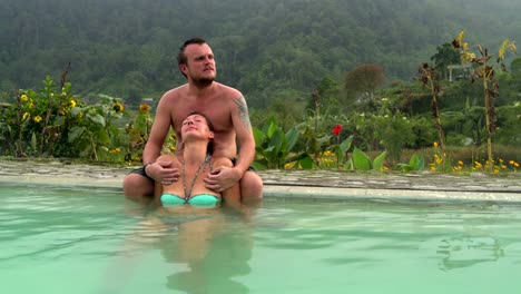 Woman-relaxes-in-a-hot-spring-with-thermal-water.-Man-sits-on-the-edge-of-the-pool-and-hugs-a-woman