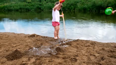 Kleine-Kinder-spielen-im-Fluss-im-Sommer,-das-Mädchen-holt-Wasser-aus-dem-Fluss-und-gießt-es-in-den-sand