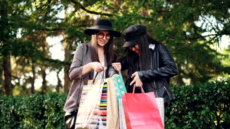 Two-young-women-shopaholics-are-talking-in-the-street-looking-at-purchases-in-bags-and-expressing-excitement.-Shopping,-people-and-happiness-concept.