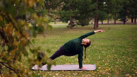 Yoga-Schüler-übt-Variationen-der-Seitenwinkel-Position-Parsvakonasana-auf-Yoga-Matte-im-Park.-Grüne-und-gelbe-Bäume,-Blätter-und-Rasen-sind-sichtbar.