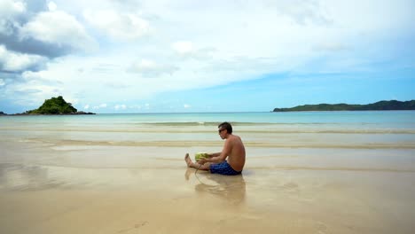 Mann-trinkt-Cocossaft-aus-einer-Mutter-an-einem-Strand-am-Meer