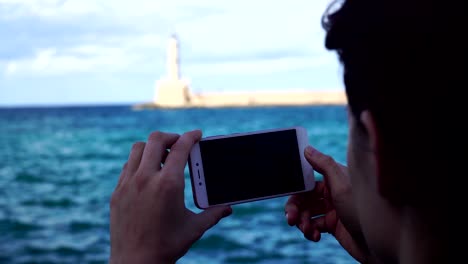 Young-woman-takes-the-photo-a-lighthouse-in-a-European-city-uses-smartphone