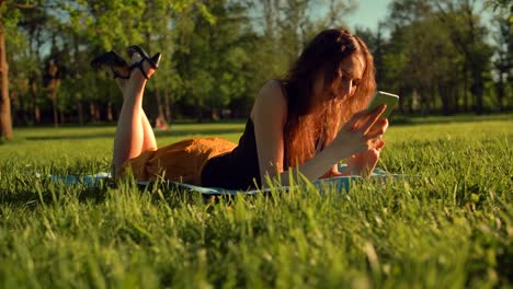 Happy-female-types-sms-or-chat,-sitting-in-park-on-grass.-Woman-chatting-in-telephone