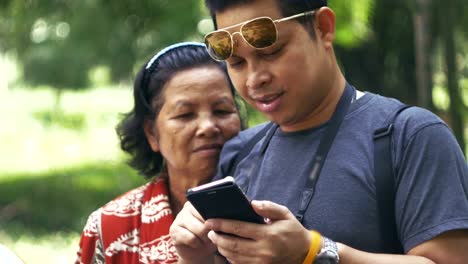 Asian-mother-and-son-rest-in-garden-while-son-using-smartphone-in-morning.