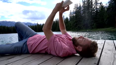 Young-man-relaxing-on-lake-pier-with-book,-Canada