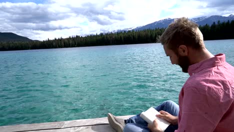 Young-man-relaxing-on-lake-pier-with-book,-Canada