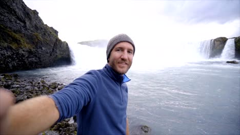 Young-man-taking-selfie-portrait-with-magnificent-waterfall-in-Iceland,-Godafoss-falls.-People-travel-exploration-concept