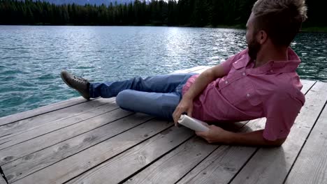 Hombre-joven-relajante-en-el-muelle-del-lago-con-el-libro,-Canadá