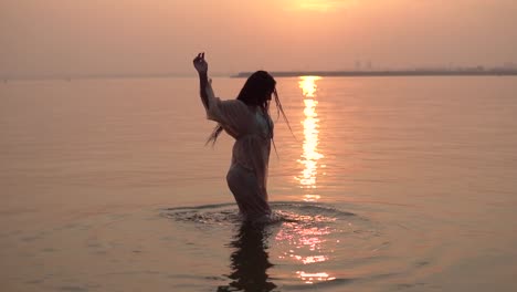 Sexy-girl-in-a-beach-tunic-dancing-standing-in-the-water-against-a-sunset-background.-slow-motion