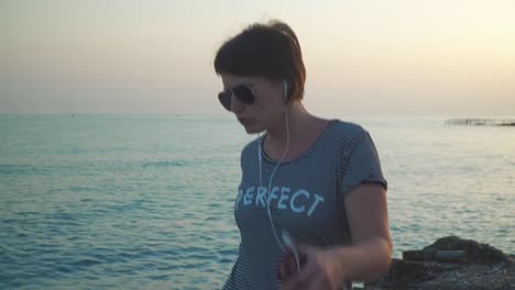 Woman-in-t-shirt-listening-to-music-in-headphones-and-dancing-in-leisure-against-sunset-seascape