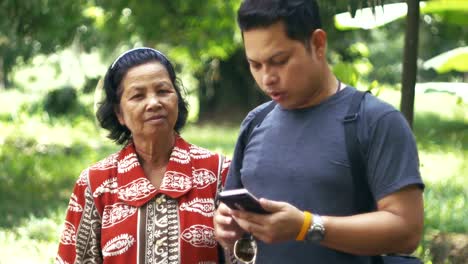 Asian-mother-and-son-rest-in-garden-while-son-using-smartphone-in-morning.