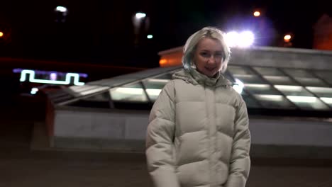 Young-blonde-woman,-in-a-white-fashion-jacket-walking-down-the-deserted-central-street-at-night