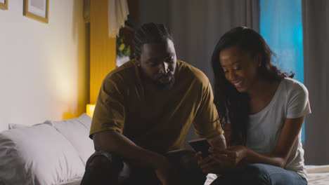Young-Couple-Relaxing-At-Home-At-Night-In-Bedroom-Looking-At-Mobile-Phone-Together-6
