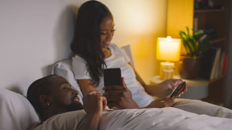 Smiling-Couple-At-Home-At-Night-Both-Looking-At-Mobile-Phones-In-Bed