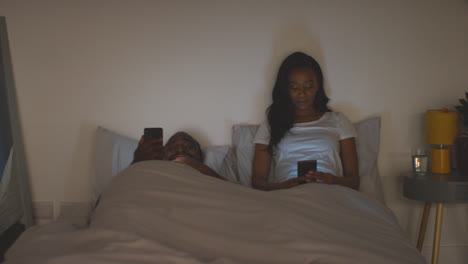 Couple-At-Home-At-Night-Both-Looking-At-Their-Mobile-Phones-In-Bed