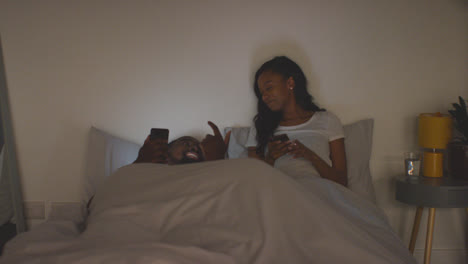 Young-Couple-Relaxing-At-Home-At-Night-In-Bed-Looking-At-Mobile-Phones-Together-1