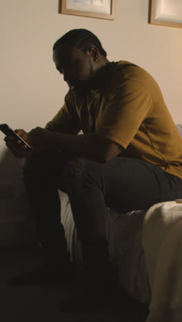 Vertical-Video-Of-Young-Man-Relaxing-At-Home-At-Night-In-Bedroom-Looking-At-Mobile-Phone