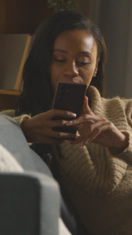 Vertical-Video-Close-Up-Of-Vertical-Video-Of-Young-Woman-Relaxing-At-Home-Sitting-On-Sofa-Scrolling-Through-Mobile-Phone