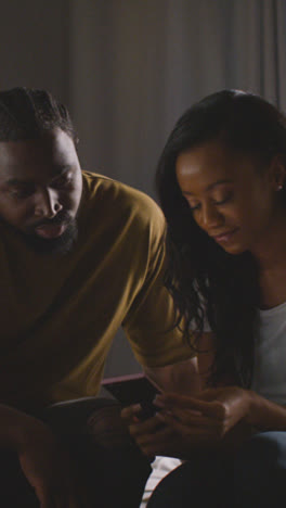 Vertical-Video-Of-Young-Couple-Relaxing-At-Home-At-Night-In-Bedroom-Looking-At-Mobile-Phone-Together