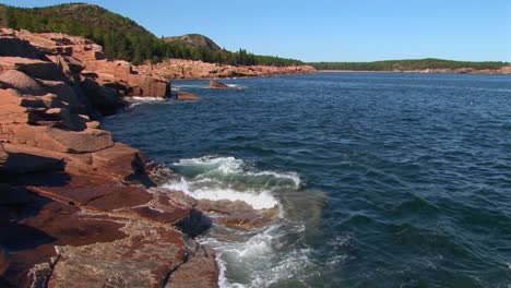 Water-crashes-against-rocks-that-are-near-trees-and-a-mountain-range-in-Acadia-National-Park-in-Maine