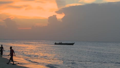 Ein-Kleines-Motorboot-Fährt-Bei-Sonnenuntergang-Heraus,-Während-Die-Leute-Am-Strand-Von-Stone-Town-Sansibar-Spazieren-Gehen
