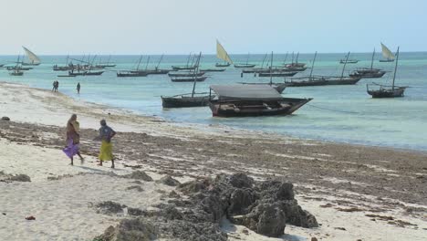 Zwei-Muslimische-Frauen-Gehen-Entlang-Eines-Strandes-In-Sansibar-Mit-Dhow-segelbooten-Im-Hintergrund