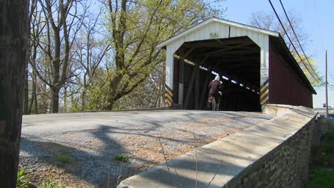 Ein-Amish-Mädchen-Fährt-Mit-Einem-Motorroller-Durch-Eine-überdachte-Brücke-Im-Ländlichen-Lancaster-Pennsylvania