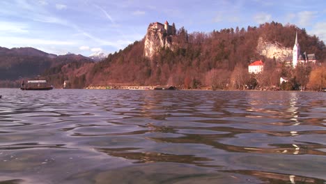 Ein-Boot-Fährt-An-Einer-Wunderschönen-Mittelalterlichen-Burg-Und-Kirche-Am-Ufer-Des-Sees-Bled-Slowenien-Vorbei
