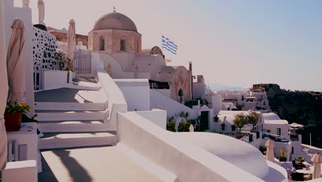 Weiße-Häuser-Säumen-Die-Hänge-Der-Griechischen-Insel-Santorini-Mit-Einer-Griechischen-Flagge-In-Der-Ferne