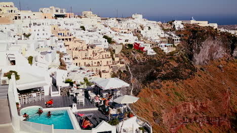 Mehrfarbige-Häuser-Säumen-Die-Hügel-Der-Griechischen-Insel-Santorini-Mit-Einer-Griechischen-Flagge-In-Der-Ferne-3