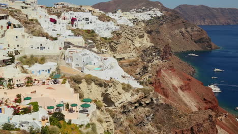 Langsame-Pfanne-Weißer-Häuser,-Die-Die-Hügel-Der-Griechischen-Insel-Santorini-Säumen,-Mit-Einer-Griechischen-Flagge-In-Der-Ferne-1
