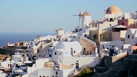 White-buildings-and-windmills-line-the-hillsides-of-the-Greek-Island-of-Santorini-with-a-Greek-flag-in-the-distance