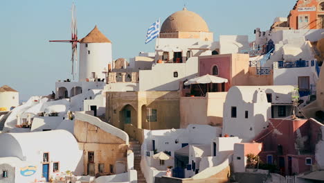 White-buildings-and-windmills-line-the-hillsides-of-the-Greek-Island-of-Santorini-with-a-Greek-flag-in-the-distance-1