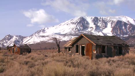 Verlassene-Siedlerhütten-Mit-Den-Schneebedeckten-Bergen-Der-Sierra-Nevada-Mit-Der-Sonne,-Die-Durch-Die-Wolken-Scheint