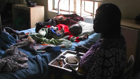 A-woman-sits-next-to-the-bed-of-a-child-in-a-hospital-and-eats-food-from-a-tray