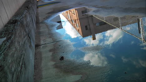 Time-lapse-of-clouds-passing-over-an-urban-building-as-reflected-in-a-puddle-on-the-pavement-