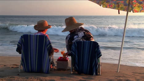 Ein-älteres-Ehepaar-Teilt-Getränke-Unter-Einem-Sonnenschirm-Am-Strand