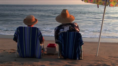 Ein-Paar-Entspannt-Sich-In-Strandkörben-Unter-Einem-Sonnenschirm-Und-Beobachtet-Das-Meer