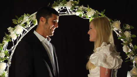 Bride-and-groom-stand-under-an-arch-of-flowers-as-the--man-slips-a-ring-on--the-woman's-finger