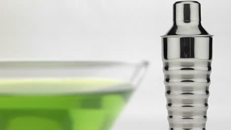 A-silver-martini-shaker-sits-behind-a-glass-with-green-liquid-inside