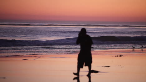Ein-Mann-Geht-Bei-Sonnenuntergang-Am-Strand-Spazieren-Und-Telefoniert-Mit-Einem-Handy