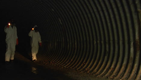 -individuals-wearing-hazmat-suits-use-flashlights-to-inspect-a-dark-tunnel