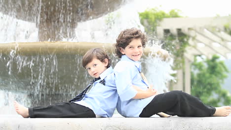 Ein-Paar-Jungen-In-Festlicher-Kleidung-Sitzen-Rücken-An-Rücken-Vor-Einem-Brunnen