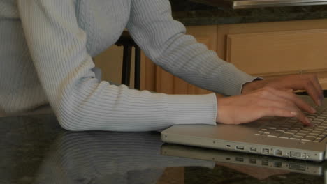 A-woman-sits-at-a-kitchen-counter-and-types-on-a-laptop-computer