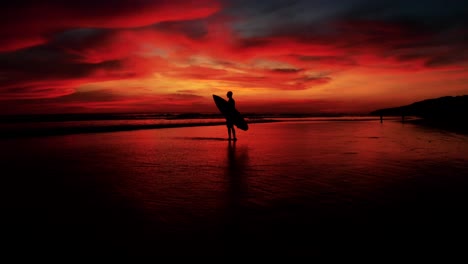 Schöne-Antenne-Eines-Surfers,-Der-Mit-Surfbrett-In-Wunderschönem-Sonnenuntergangsrotlicht-Steht